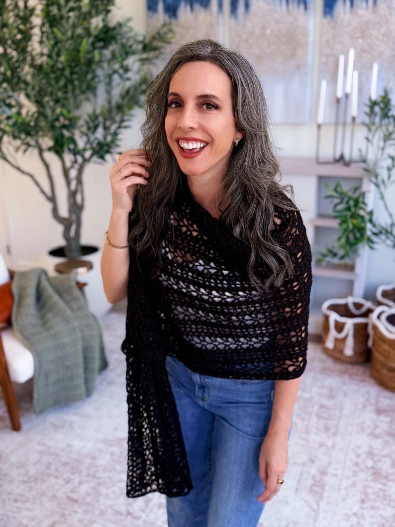 May include: A black crocheted shawl with an openwork pattern draped over the shoulders, paired with blue denim jeans and a white top. The shawl features a repeating geometric stitch design and hangs down on one side, showcasing the intricate texture.