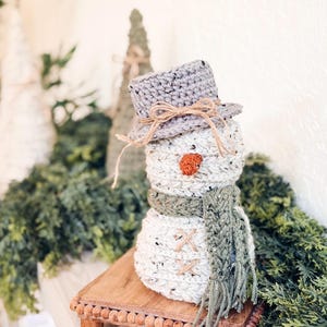 Speckled cream crochet snowman in gray hat and green scarf on a wooden riser near crochet trees and greenery