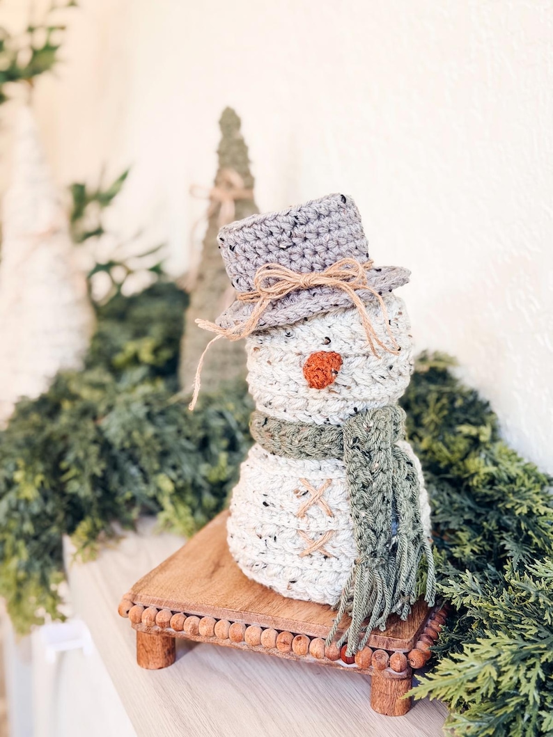Speckled cream crochet snowman in gray hat and green scarf on a wooden riser near crochet trees and greenery