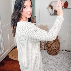 A woman in a white crochet sweater, adorned with unique jewellery, is posing and smiling at the camera as she appears to take a selfie with her smartphone in a well-lit room with minimalist decor