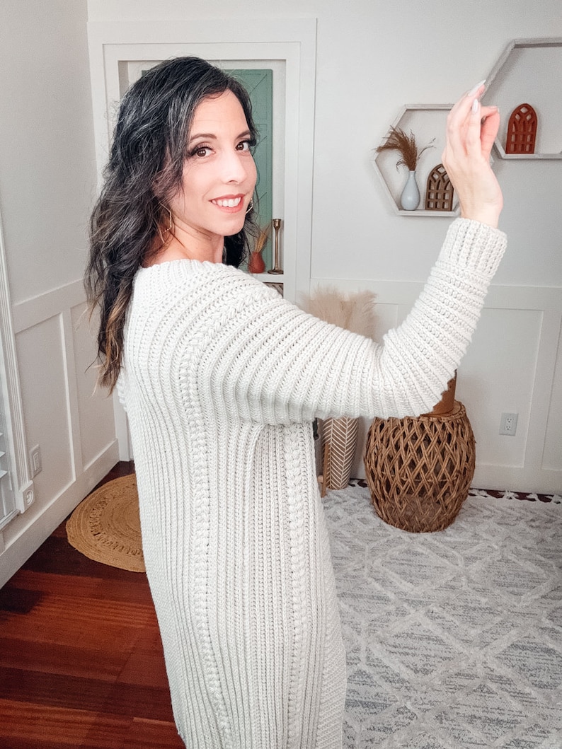 A woman in a white crochet sweater, adorned with unique jewellery, is posing and smiling at the camera as she appears to take a selfie with her smartphone in a well-lit room with minimalist decor