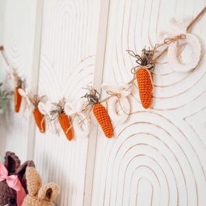 May include: An Easter garland with crocheted orange carrots and white bunny shapes, strung on twine. The garland hangs against white wooden panels with carved arch patterns. Crochet bunnies in the foreground add to the Easter theme. Carrots are approximately 7 cm long.