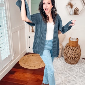 A woman in a white top, blue jeans, and a crochet cardigan poses playfully with one hand on her hip and the other extended to the side, wearing unique jewellery, standing in a room