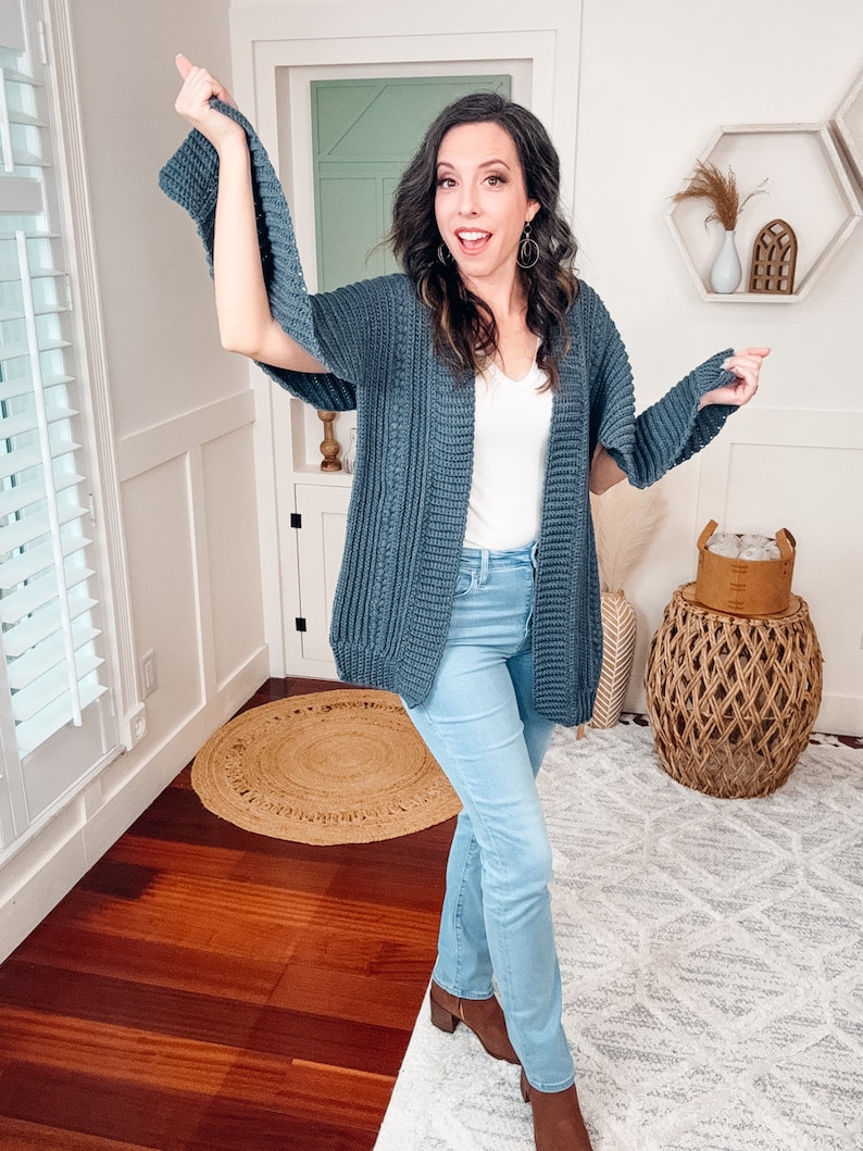 A woman in a white top, blue jeans, and a crochet cardigan poses playfully with one hand on her hip and the other extended to the side, wearing unique jewellery, standing in a room