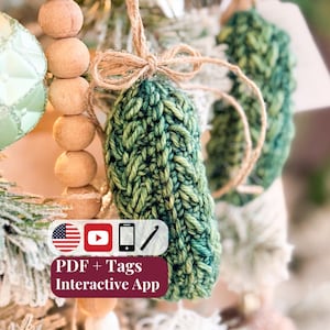 May include: A green crocheted ornament with a textured stitch pattern, tied with a natural jute twine bow. A rectangular label displays the text: PDF + Tags Interactive App, accompanied by icons of a US flag, YouTube play button, phone, and crochet hook.
