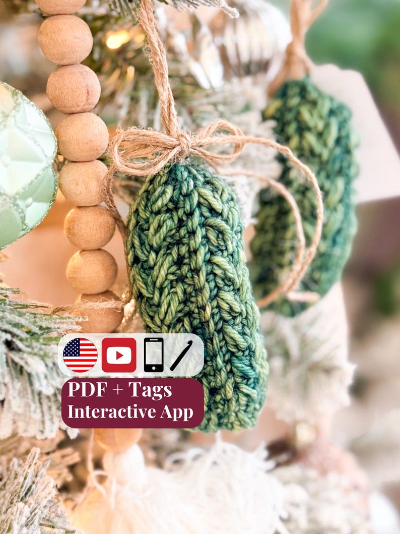 May include: A green crocheted ornament with a textured stitch pattern, tied with a natural jute twine bow. A rectangular label displays the text: PDF + Tags Interactive App, accompanied by icons of a US flag, YouTube play button, phone, and crochet hook.