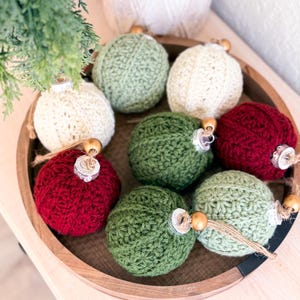 May include: A collection of seven spherical baubles covered in crocheted yarn in shades of cream, sage green, forest green, and deep red. Each bauble features a silver-toned cap, a natural jute twine loop, and a small wooden bead accent.
