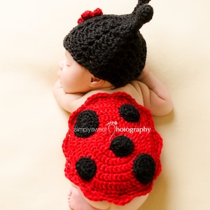 May include: A baby wearing a black and red ladybug costume. The costume is crocheted and has black dots on a red background. The baby is lying on its side with its head resting on its arm.