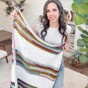 Puede incluir: Una manta de ganchillo hecha a mano con una base blanca y rayas coloridas en turquesa, morado, dorado y rosa. La manta se sostiene, mostrando su textura y diseño. Un elemento de decoración del hogar acogedor.