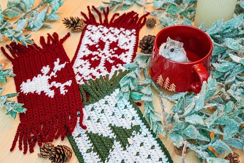 May include: Two crocheted scarves with a snowflake and reindeer design in red, white, and green. A small hamster is sitting in a red mug with a tree design.