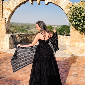 May include: A black crochet shawl with an openwork pattern draped over the shoulders, paired with a black sleeveless top and a long, flowing black skirt. The shawl features a rectangular shape with a detailed, repeating stitch design throughout.