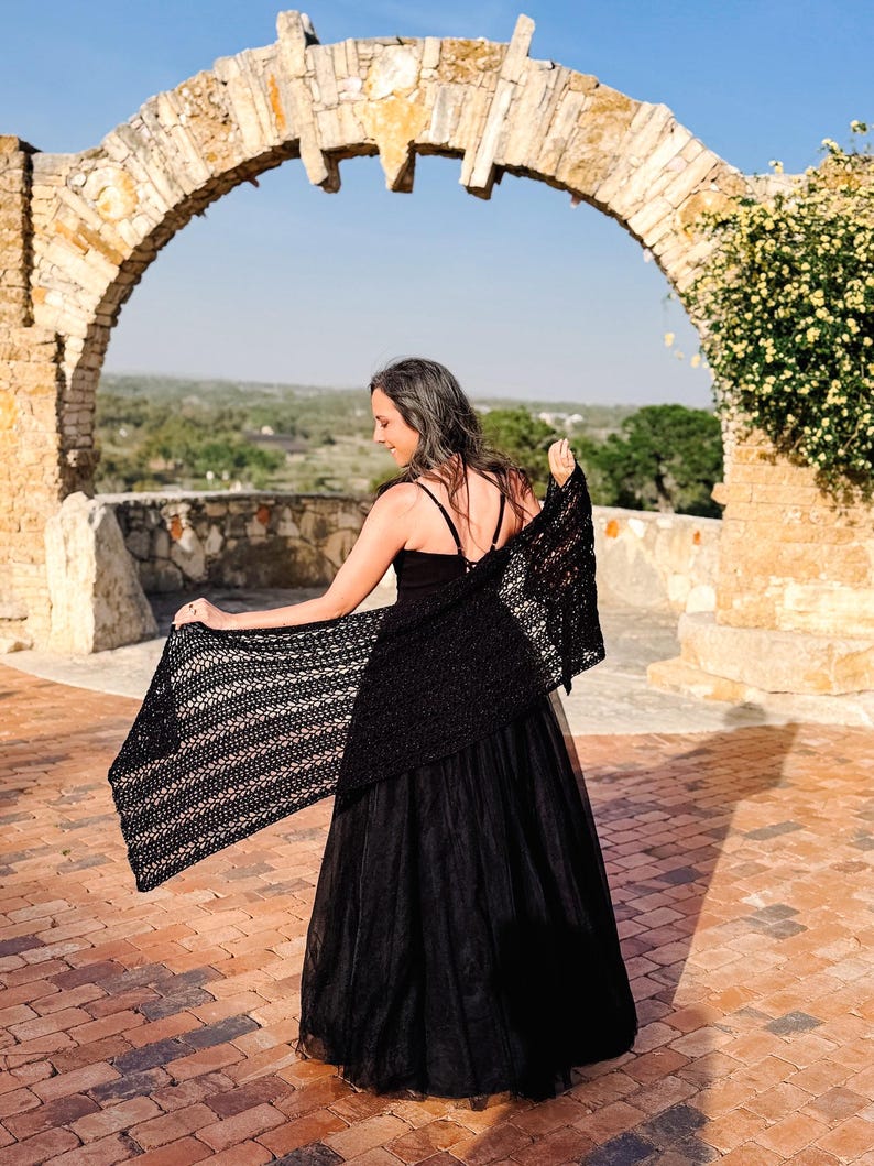 May include: A black crochet shawl with an openwork pattern draped over the shoulders, paired with a black sleeveless top and a long, flowing black skirt. The shawl features a rectangular shape with a detailed, repeating stitch design throughout.