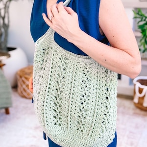Puede incluir: Un bolso tote de ganchillo verde claro, de tejido abierto, con una correa para el hombro. El bolso tiene un fondo redondeado y un patr&oacute;n texturizado. La persona de la imagen lleva una blusa y pantalones azules. El bolso es de color verde claro.