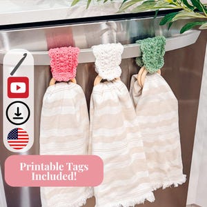 May include: Three decorative kitchen towels hanging on a stainless steel appliance. Each towel has a crocheted topper in pink, white, and green, attached to a wooden ring. The towels are beige with white stripes. The image includes the text "Printable Tags Included!"