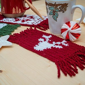 May include: Red, green, and white crocheted coasters with fringe, featuring a reindeer design. The coasters are on a wooden surface with a white star and a red and white candy cane ornament.