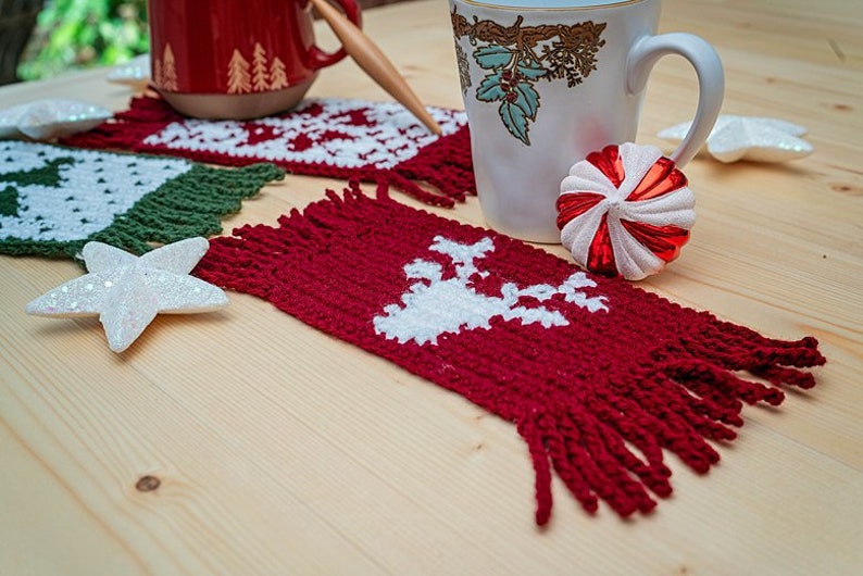 May include: Red, green, and white crocheted coasters with fringe, featuring a reindeer design. The coasters are on a wooden surface with a white star and a red and white candy cane ornament.