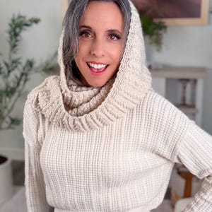May include: Cream-coloured knit sweater with a matching hood and cowl neck. The sweater has a ribbed texture and long sleeves. The person is wearing blue jeans. The sweater is a cosy and stylish garment.