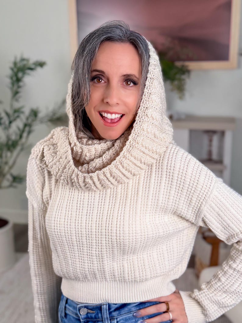 May include: Cream-coloured knit sweater with a matching hood and cowl neck. The sweater has a ribbed texture and long sleeves. The person is wearing blue jeans. The sweater is a cosy and stylish garment.