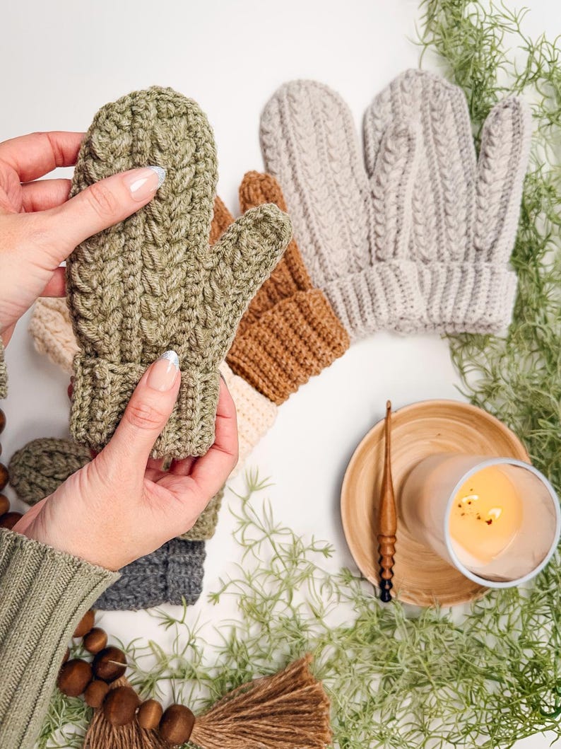 May include: A set of five crocheted mittens in various colours, including green, brown, beige, and grey. The mittens are displayed on a white surface with green foliage and a wooden candle holder.
