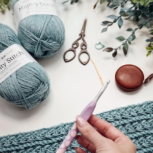 May include: A craft scene featuring teal yarn balls labeled "Mighty Stitch", vintage scissors, a crochet hook, and a brown leather tape measure. A hand holds the crochet hook over a teal crocheted fabric.