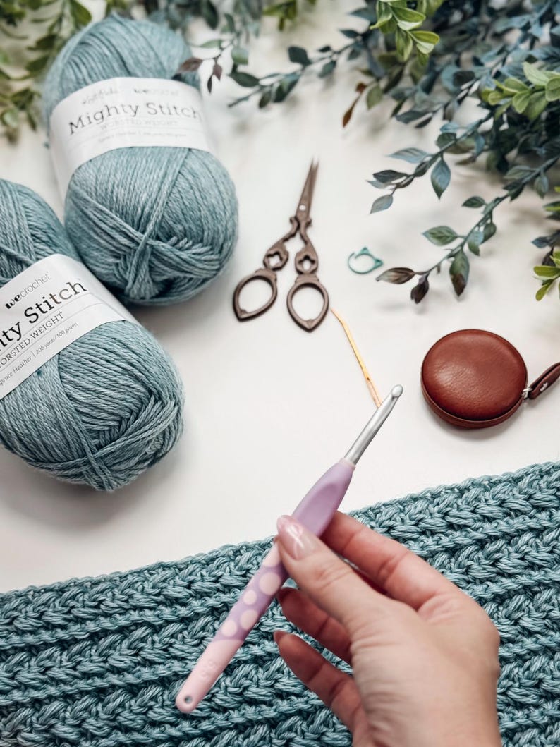 May include: A craft scene featuring teal yarn balls labeled "Mighty Stitch", vintage scissors, a crochet hook, and a brown leather tape measure. A hand holds the crochet hook over a teal crocheted fabric.