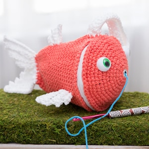 May include: A coral-colored crocheted fish-shaped pouch featuring a white zipper closure, white crocheted fins, and large googly eyes. A blue yarn tail extends from the fish's mouth, resting near a metal crochet hook with a patterned handle.