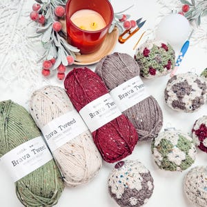 May include: Four skeins of Brava Tweed worsted weight yarn in olive green, cream, cranberry red, and heather grey, each with a white label. Beside them are several crocheted spherical ornaments in matching colours, a red candle, and a crochet hook.