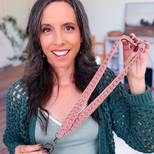May include: A dusty rose-colored, braided lanyard with a silver-tone swivel clasp. The lanyard is held up by a person. The person is wearing a green crochet cardigan and a light green tank top. A set of keys is attached to the lanyard.