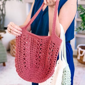 Puede incluir: Un bolso tote de ganchillo rosa polvoriento con asas largas. El bolso tiene un dise&ntilde;o abierto y de encaje y lo sostiene una persona. Tambi&eacute;n se ven otros dos bolsos de ganchillo, uno blanco y otro verde claro.