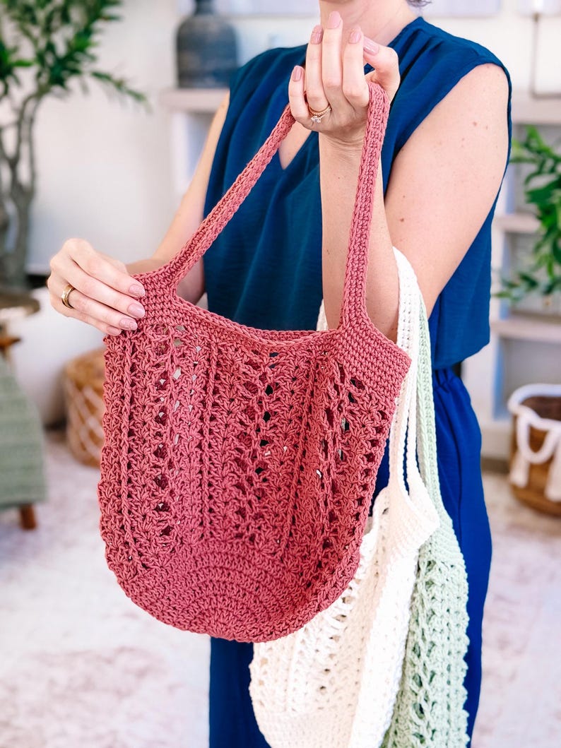 Puede incluir: Un bolso tote de ganchillo rosa polvoriento con asas largas. El bolso tiene un dise&ntilde;o abierto y de encaje y lo sostiene una persona. Tambi&eacute;n se ven otros dos bolsos de ganchillo, uno blanco y otro verde claro.