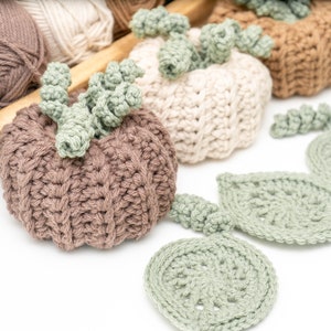 Taupe and cream crochet pumpkins with sage leaf coasters spread out beside yarn skeins in a wooden tray