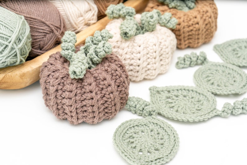 Taupe and cream crochet pumpkins with sage leaf coasters spread out beside yarn skeins in a wooden tray
