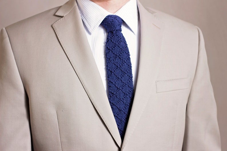 May include: A beige suit jacket with a blue knitted tie. The jacket has a single button closure and a pocket on the left side.