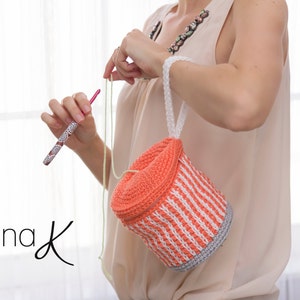 Pode incluir: Uma mulher está a fazer crochet numa bolsa de crochet de cor laranja brilhante e branca com uma alça branca. A bolsa é de forma cilíndrica e tem uma guarnição de crochet cinzenta em torno da parte inferior.