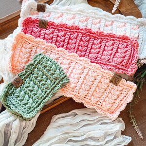 May include: A collection of handmade crochet headbands in various colors, including white, pink, peach, and green. Each headband features a unique textured stitch pattern and a small leather tag. The headbands are displayed on a wooden tray with a white fabric.