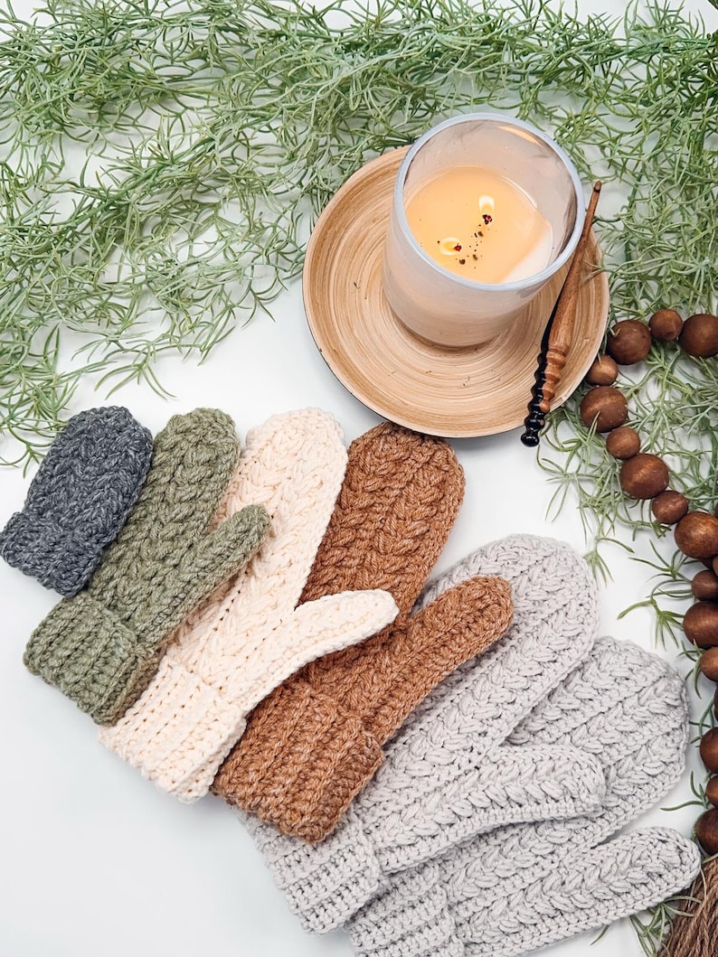 May include: A set of six crocheted mittens in various colours, including grey, green, beige, brown, and white. The mittens are arranged in a row on a white surface.