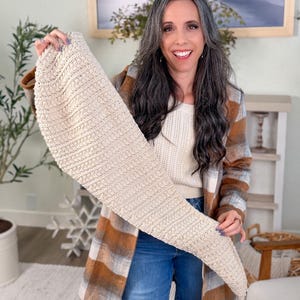 May include: A beige, knitted scarf with a cable knit pattern is displayed. The scarf is a light neutral colour. The person in the image is wearing a plaid jacket with brown and white tones, blue jeans, and a white sweater.