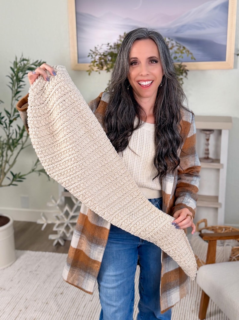 May include: A beige, knitted scarf with a cable knit pattern is displayed. The scarf is a light neutral colour. The person in the image is wearing a plaid jacket with brown and white tones, blue jeans, and a white sweater.