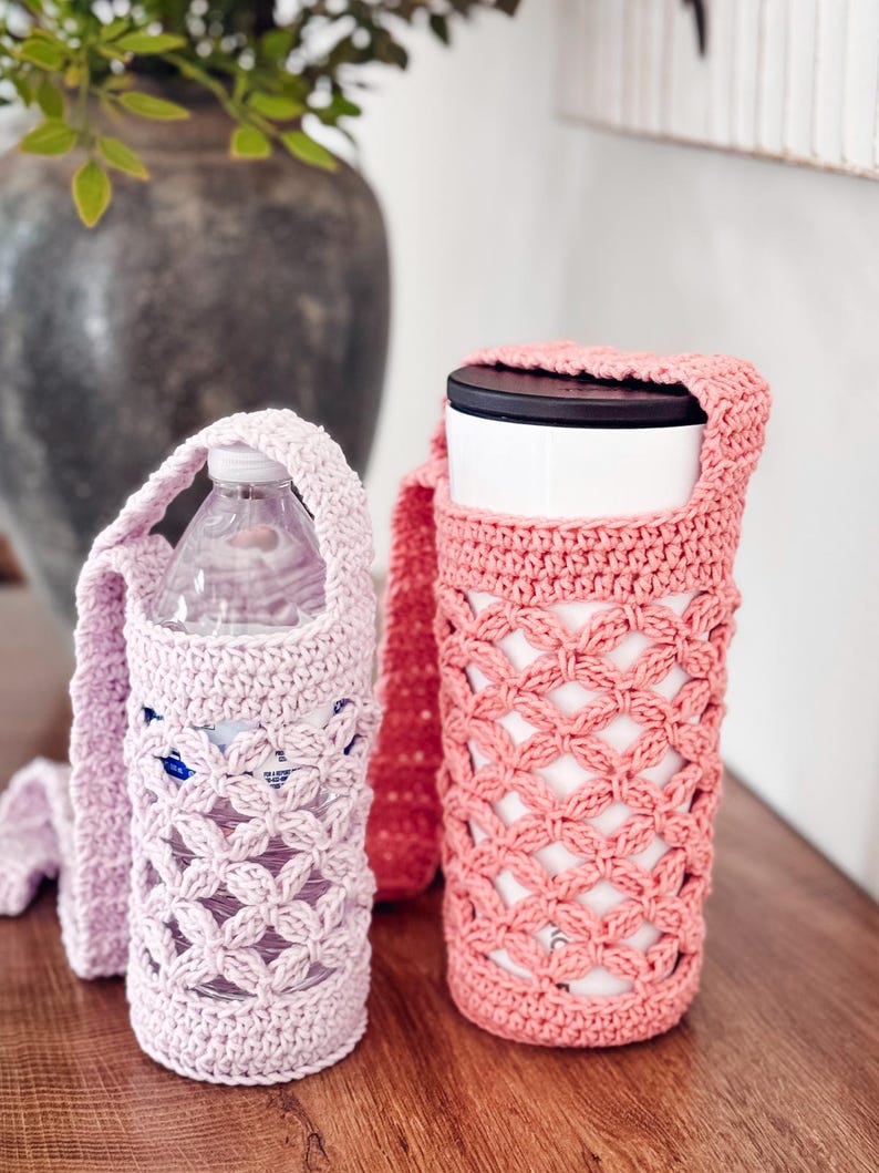 Puede incluir: Dos portabotellas de ganchillo con correas largas. El soporte de la izquierda es de color rosa claro y contiene una botella de agua de pl&aacute;stico transparente. El de la derecha es de color rosa coral y contiene un vaso blanco con tapa negra.