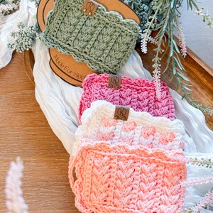 May include: A collection of handmade crochet cup cozies in various colors, including sage green, pink, white, and peach. Each cozy features a textured stitch pattern and a scalloped edge. Small leather tags are attached to each cozy.