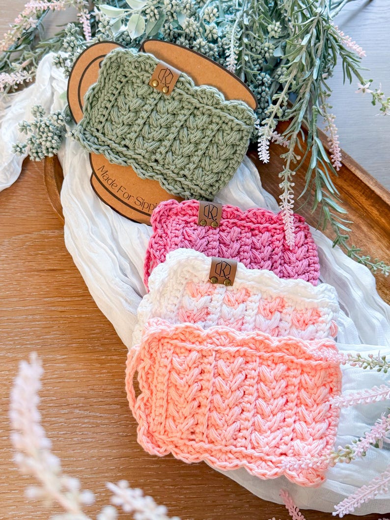May include: A collection of handmade crochet cup cozies in various colors, including sage green, pink, white, and peach. Each cozy features a textured stitch pattern and a scalloped edge. Small leather tags are attached to each cozy.