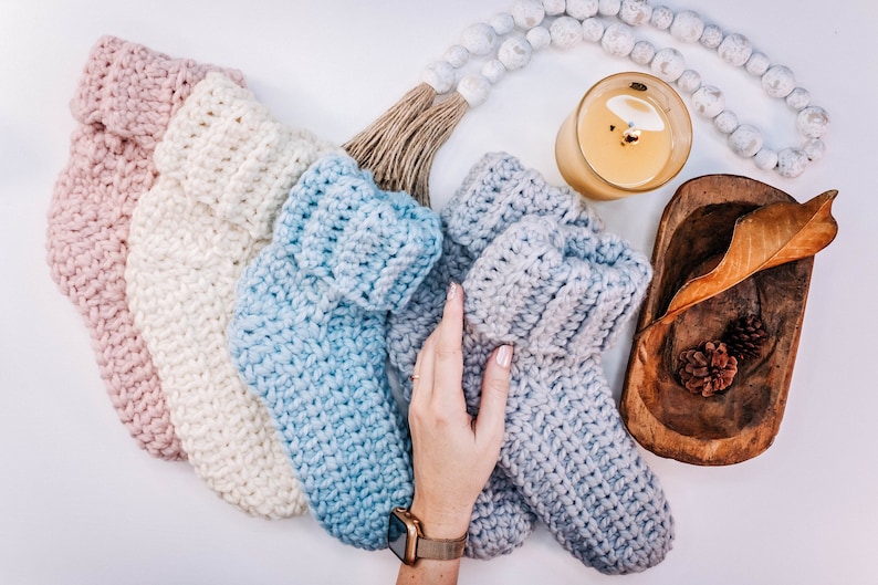 May include: Four pairs of crocheted slipper socks in various shades of pink, white, blue, and grey. The socks are arranged in a semi-circle on a white surface.