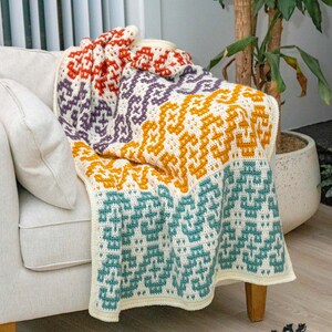 Puede incluir: Una manta de ganchillo colorida con un patr&oacute;n geom&eacute;trico en tonos naranja, amarillo, morado y turquesa. La manta est&aacute; drapeada sobre un sill&oacute;n blanco y una alfombra de color crema con flecos negros.