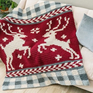 Puede incluir: Una manta de punto de crochet roja, blanca y gris con un patrón de renos. La manta está drapeada sobre un sofá blanco con almohadas grises.