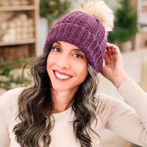 Purple crochet beanie with textured stitches and faux fur pom pom worn by a smiling woman