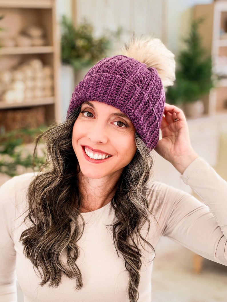 Purple crochet beanie with textured stitches and faux fur pom pom worn by a smiling woman