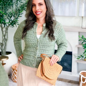 May include: A light green crochet cardigan with button closures. The cardigan is paired with a beige skirt and a straw handbag with a brown leather handle. The woman is smiling and standing in front of a fireplace.