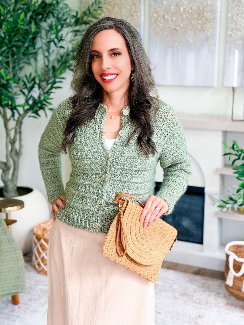 May include: A light green crochet cardigan with button closures. The cardigan is paired with a beige skirt and a straw handbag with a brown leather handle. The woman is smiling and standing in front of a fireplace.