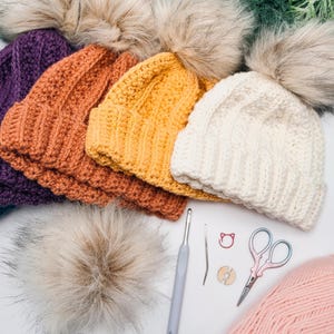Purple, orange, yellow and cream crochet beanies with fur pom poms beside a hook, scissors and pink yarn
