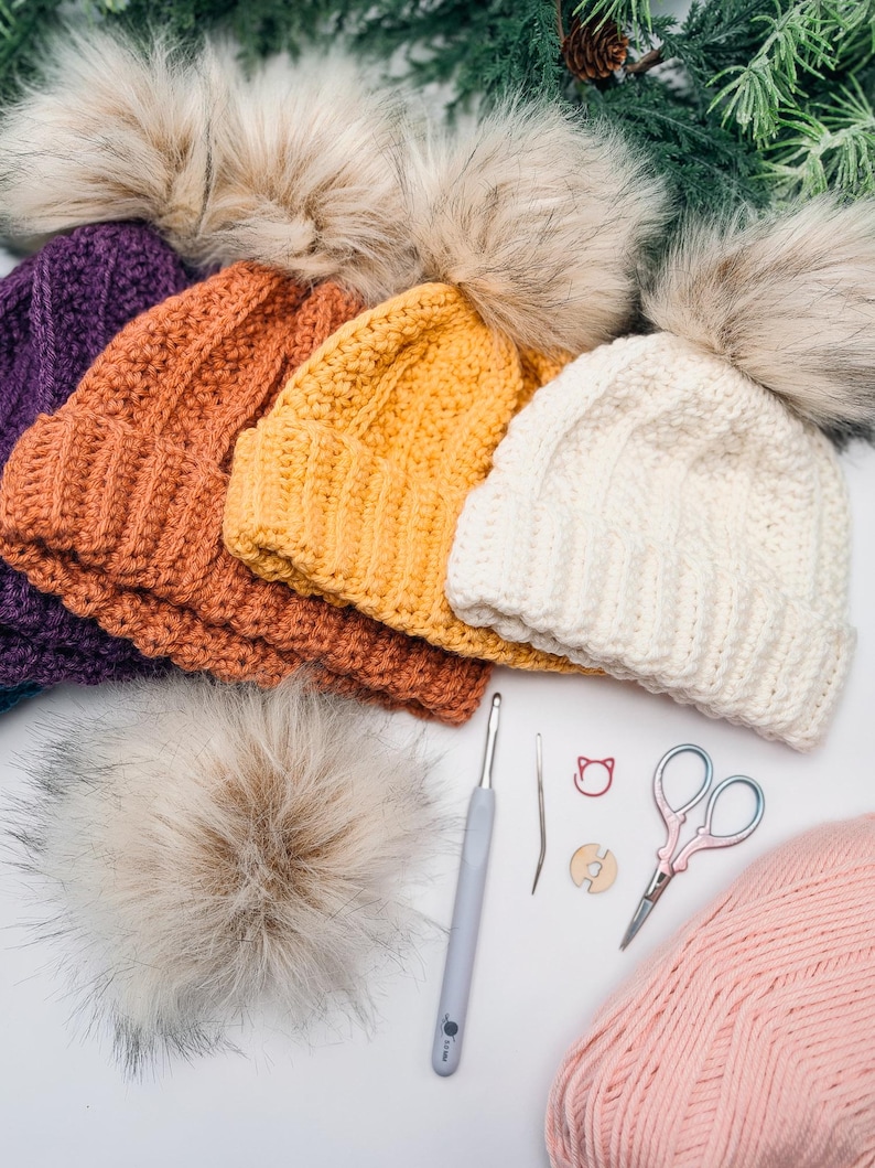 Purple, orange, yellow and cream crochet beanies with fur pom poms beside a hook, scissors and pink yarn